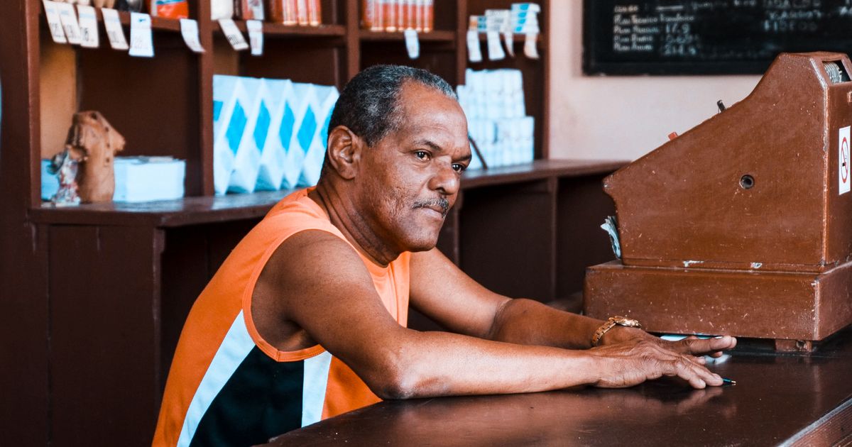 Comerciante apoiado no balcão de madeira de uma loja, ao lado da caixa registradora