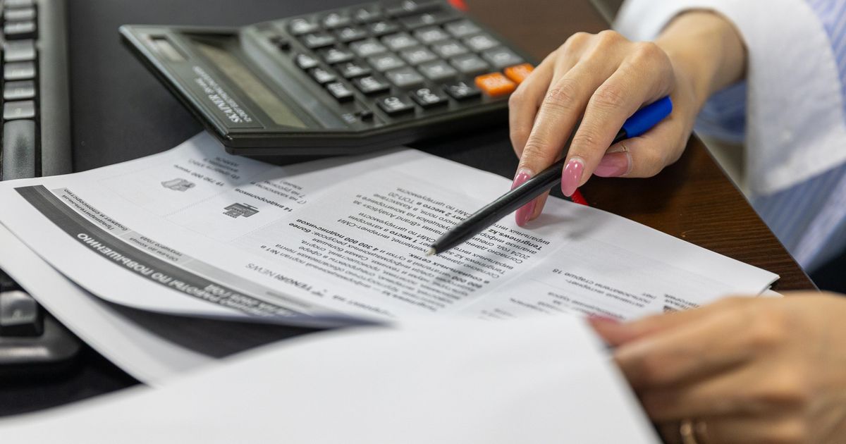 Mão de uma mulher revisando documentos financeiros ao lado de uma calculadora